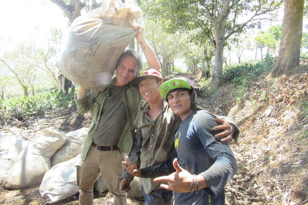 Drei Personen auf einer Kaffeeplantage. Die Person links trägt einen Kaffeesack auf der Schulter